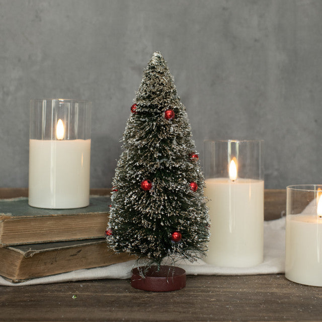 A small artificial green bottlebrush tree with red berries on a table, accompanied by candles and books.