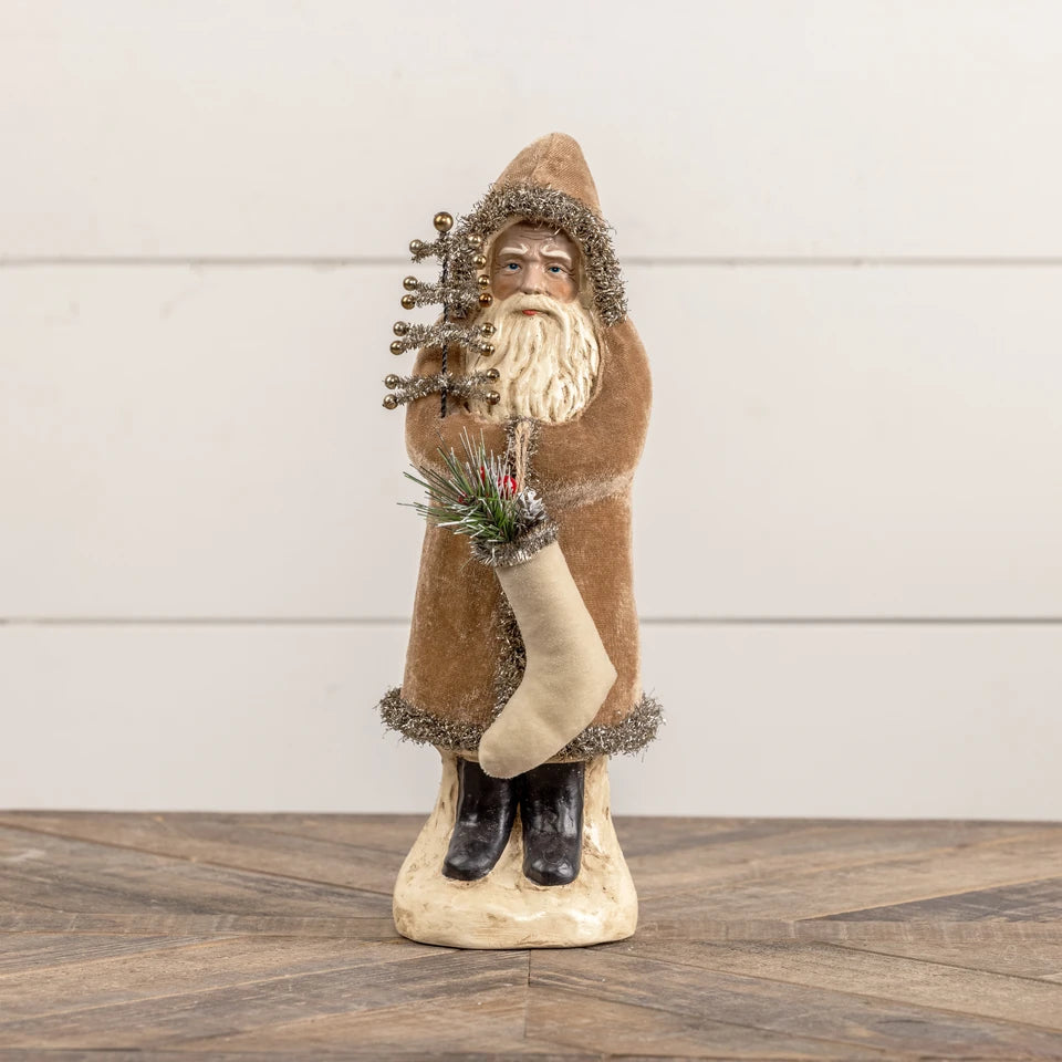 A Santa Claus holiday figurine wearing a brown fur-trimmed robe and holding a stocking and a small tree, displayed on a wooden surface against a white paneled wall.