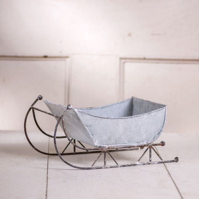 A 13-inch tin sleigh with a rustic white wash finish and solid steel runners, displayed on a hearth or tabletop.