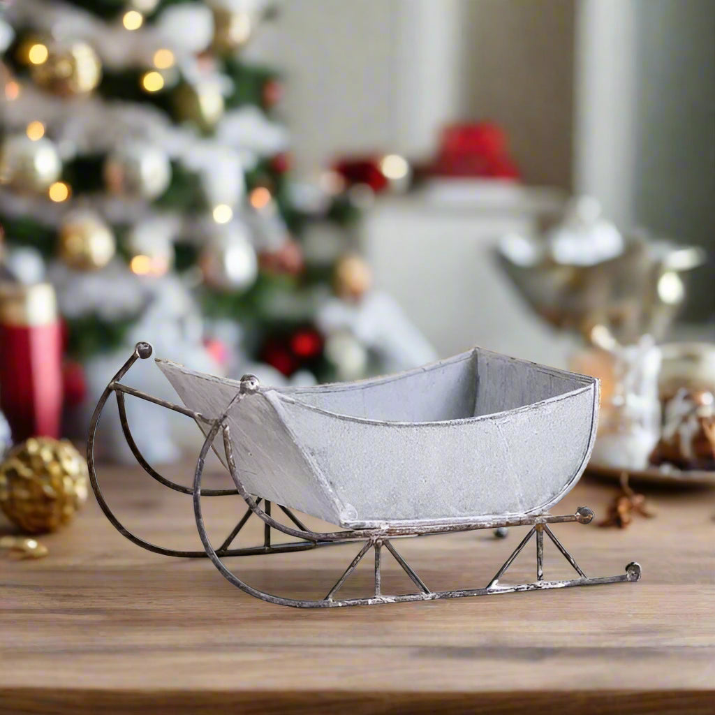 A 13-inch tin sleigh with a rustic white wash finish and solid steel runners, displayed on a hearth or tabletop.