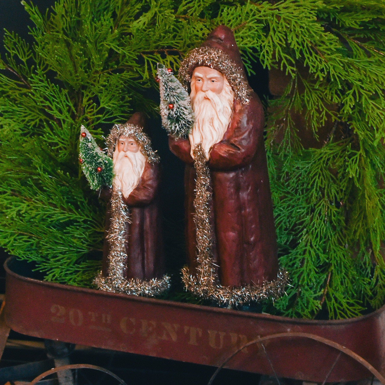 Two red belsnickels holding bottlebrush trees stand in old wagon with green wreath behind them.