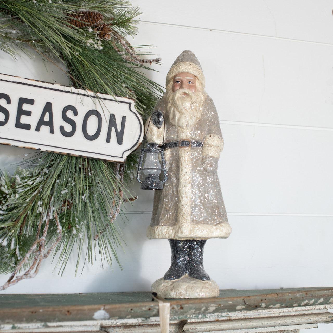 A taupe-colored belsnickel figurine holding a lantern, standing in front of a 'SEASON' sign, with holiday decorations in the background.