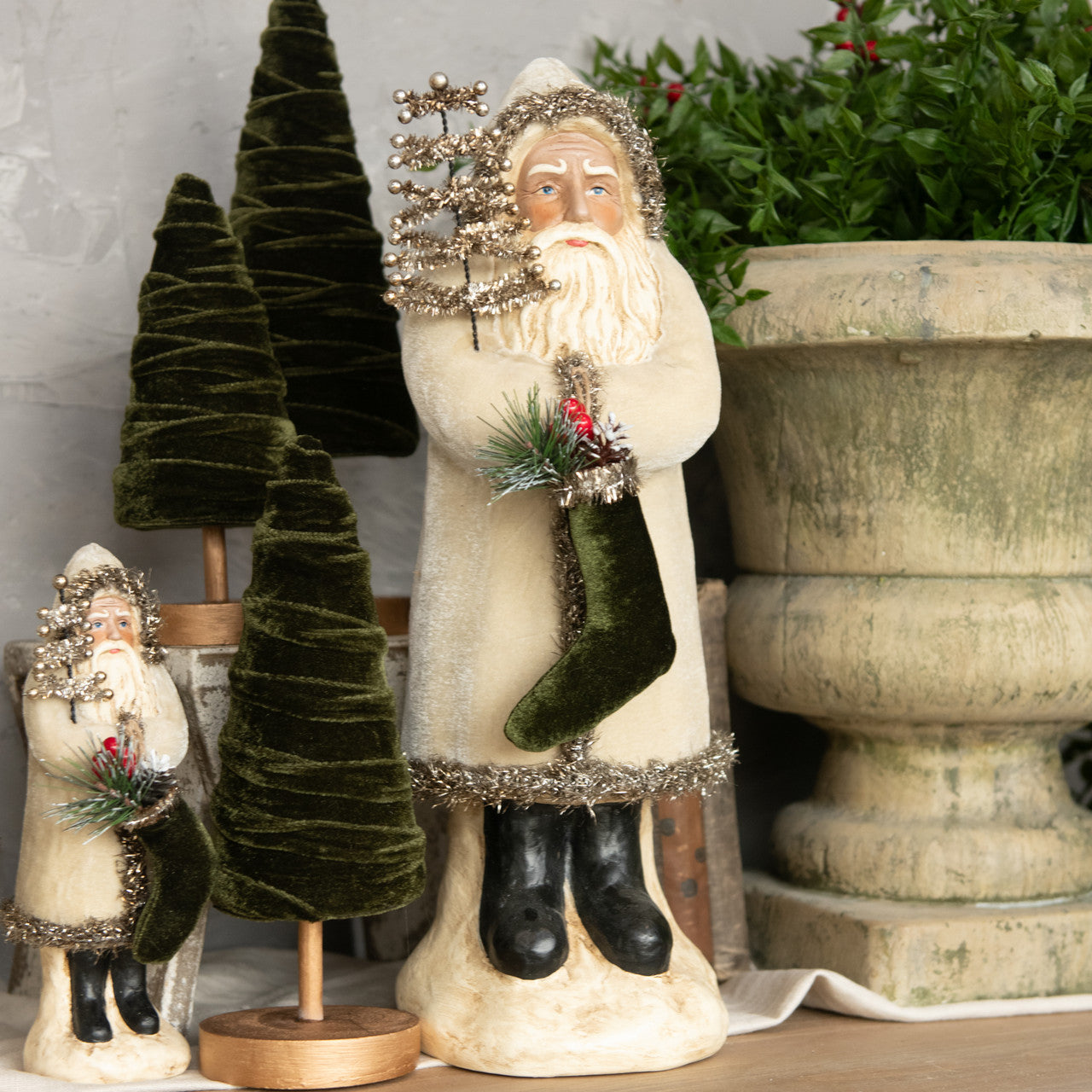 A pair of belsnickle figurines made of oatmeal velvet, one large and one small, with the large one holding a Santa hat, standing beside a decorative potted plant and a wooden object, all part of a holiday decoration setup.