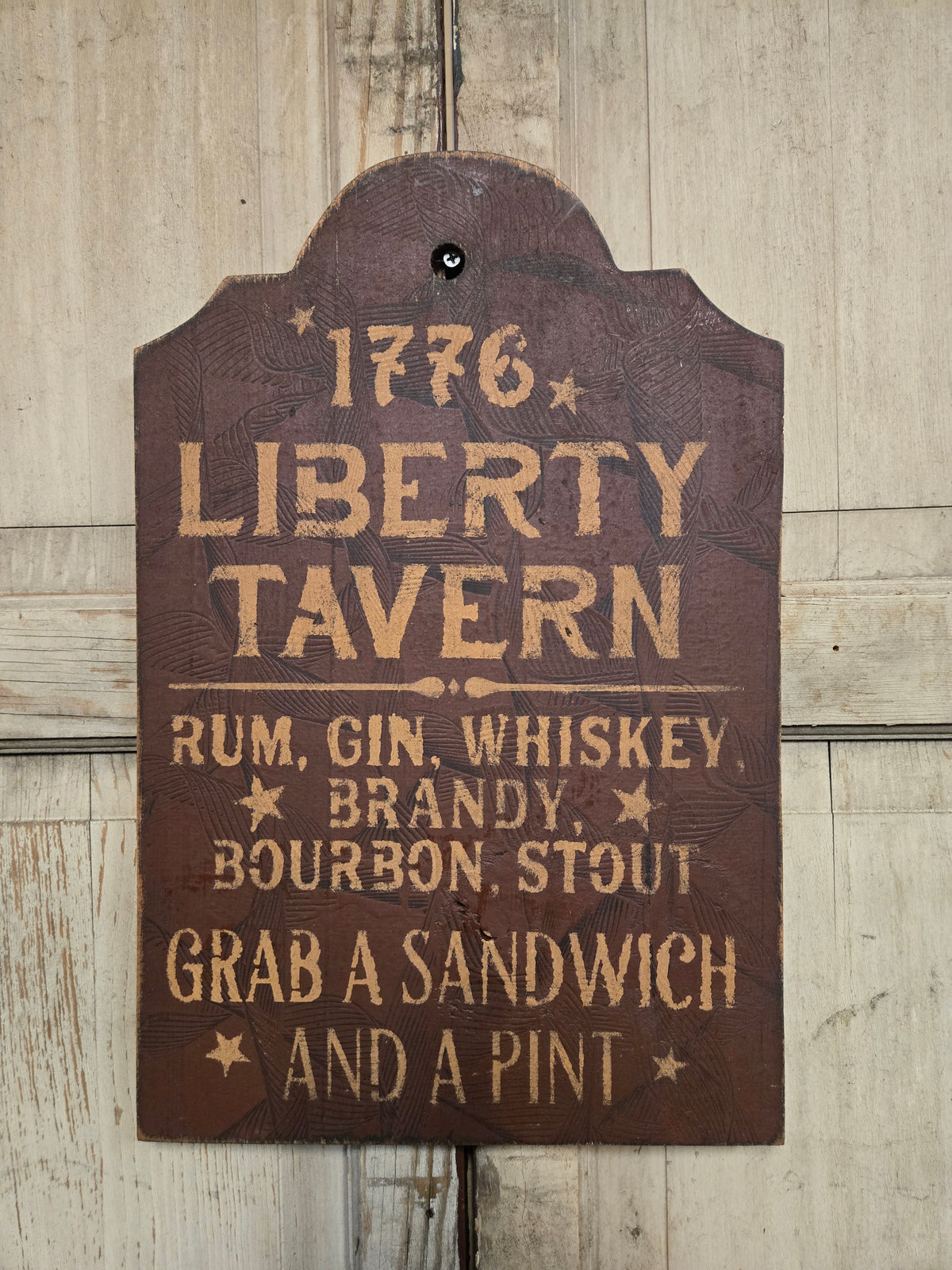 A rustic brown wooden sign with a scalloped top edge hanging on a light wood plank wall. The sign features distressed tan lettering that reads '1776 LIBERTY TAVERN' and 'RUM, GIN, WHISKEY, BRANDY, BOURBON STOUT' above 'GRAB A SANDWICH AND A PINT'.