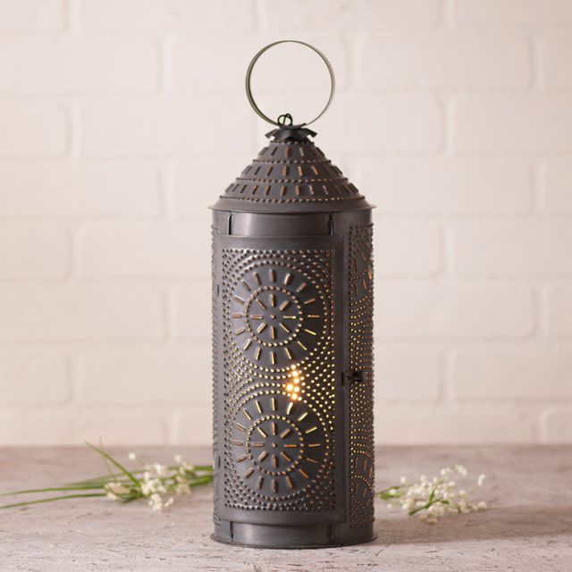 An 18-inch black chimney lantern with a geometric punch pattern, featuring a candle inside, placed on a counter against a white brick wall background.