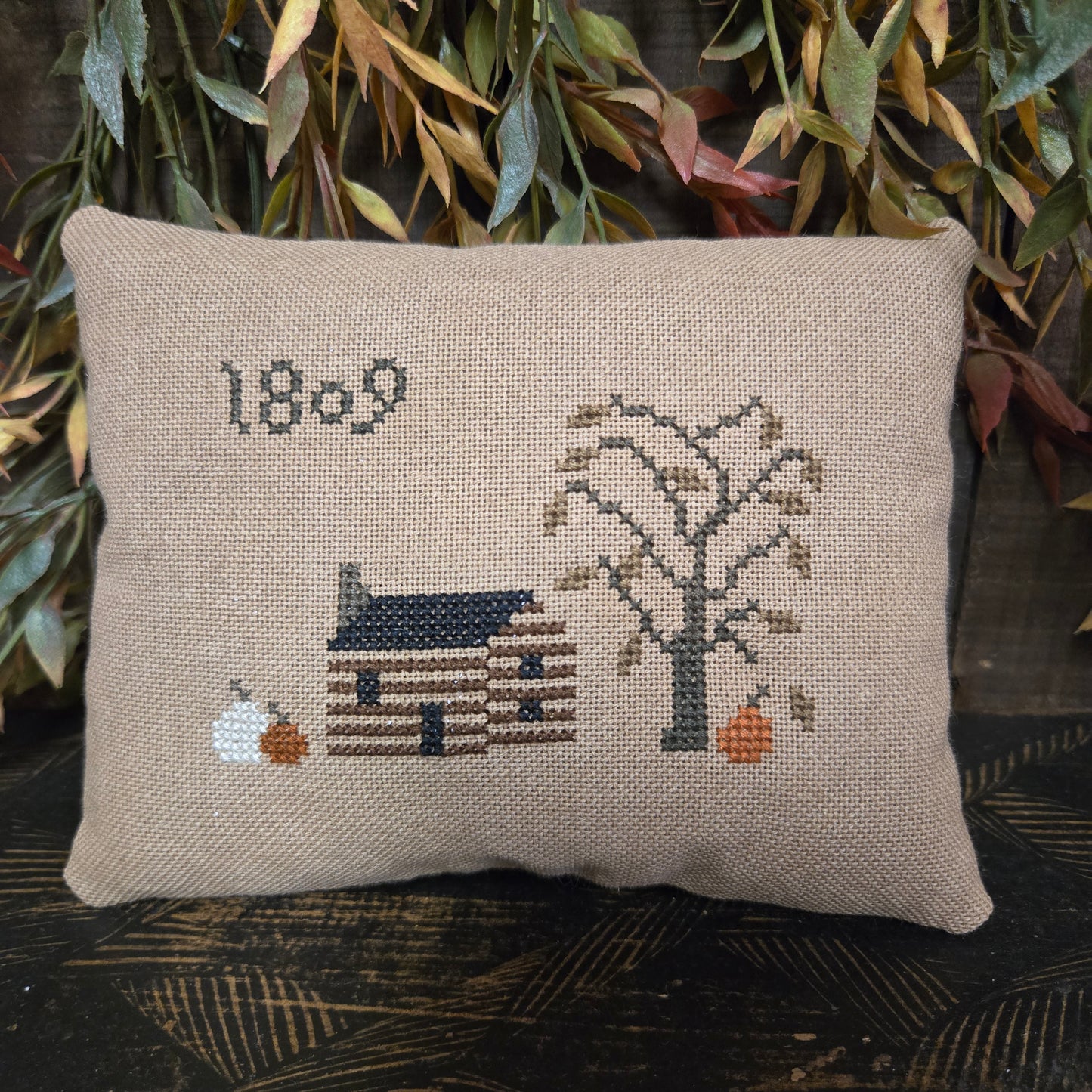 Decorative pillow with cross-stitched design of a house and tree on a wooden surface.