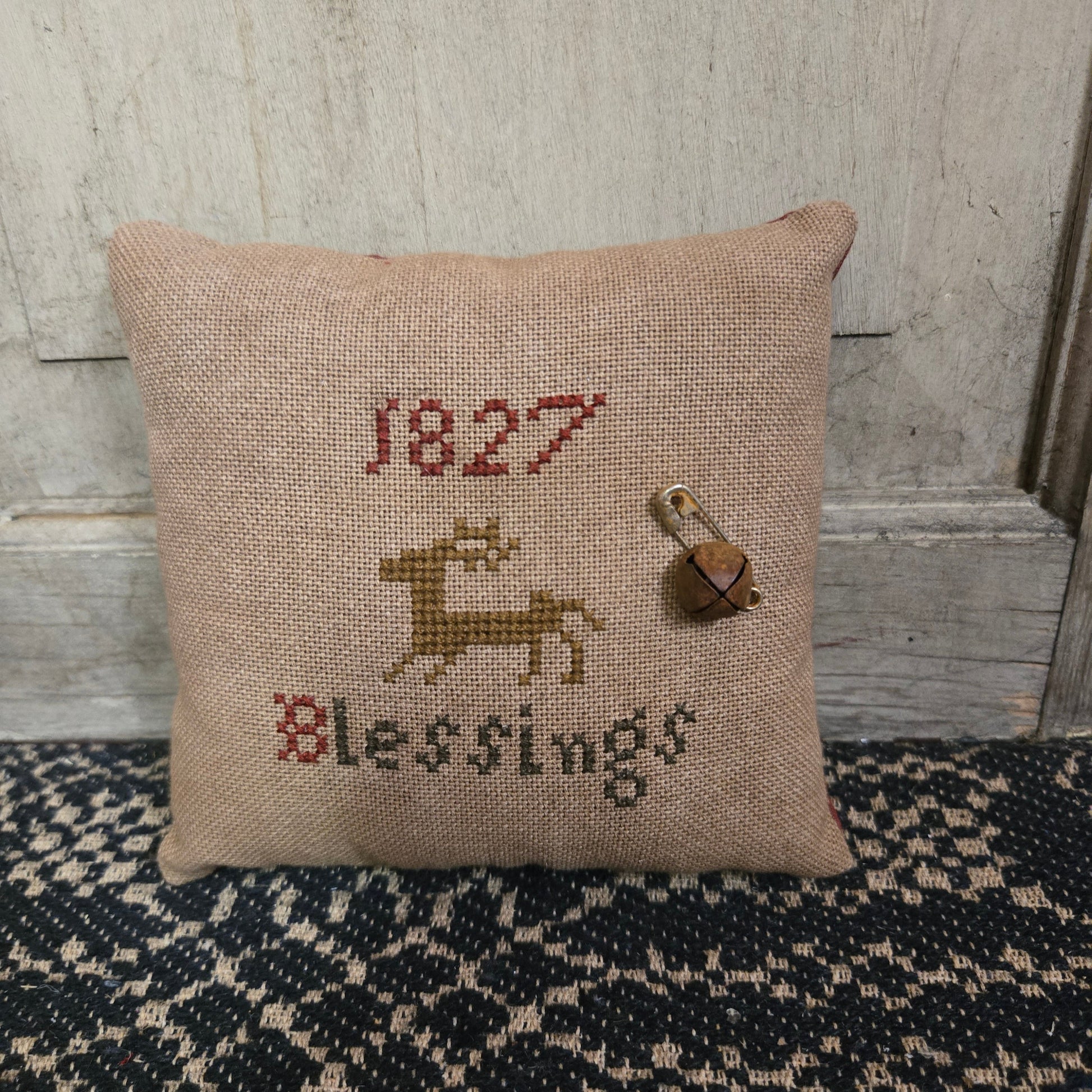 A square cross stitch pillow with the text '1827 Blessings' and a reindeer motif, displayed against a wooden background.