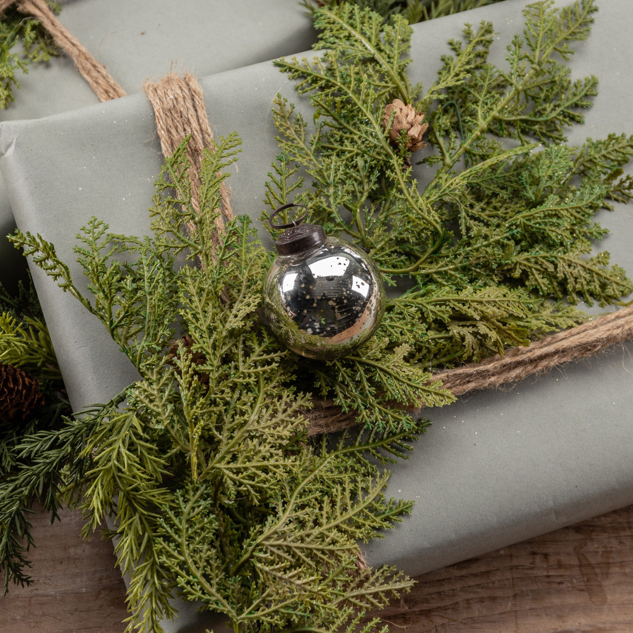A 2-inch silver kugel ornament placed on a bed of green pine needles, with a burlap bow on a gift wrapped in a neutral-colored paper.
