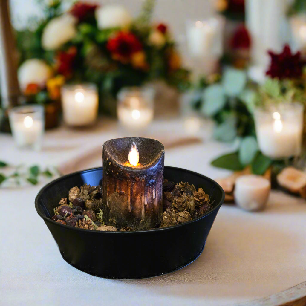 A black moving flame pillar candle on a rustic metal dish with a flickering light, placed on a patterned mat against a wooden background.