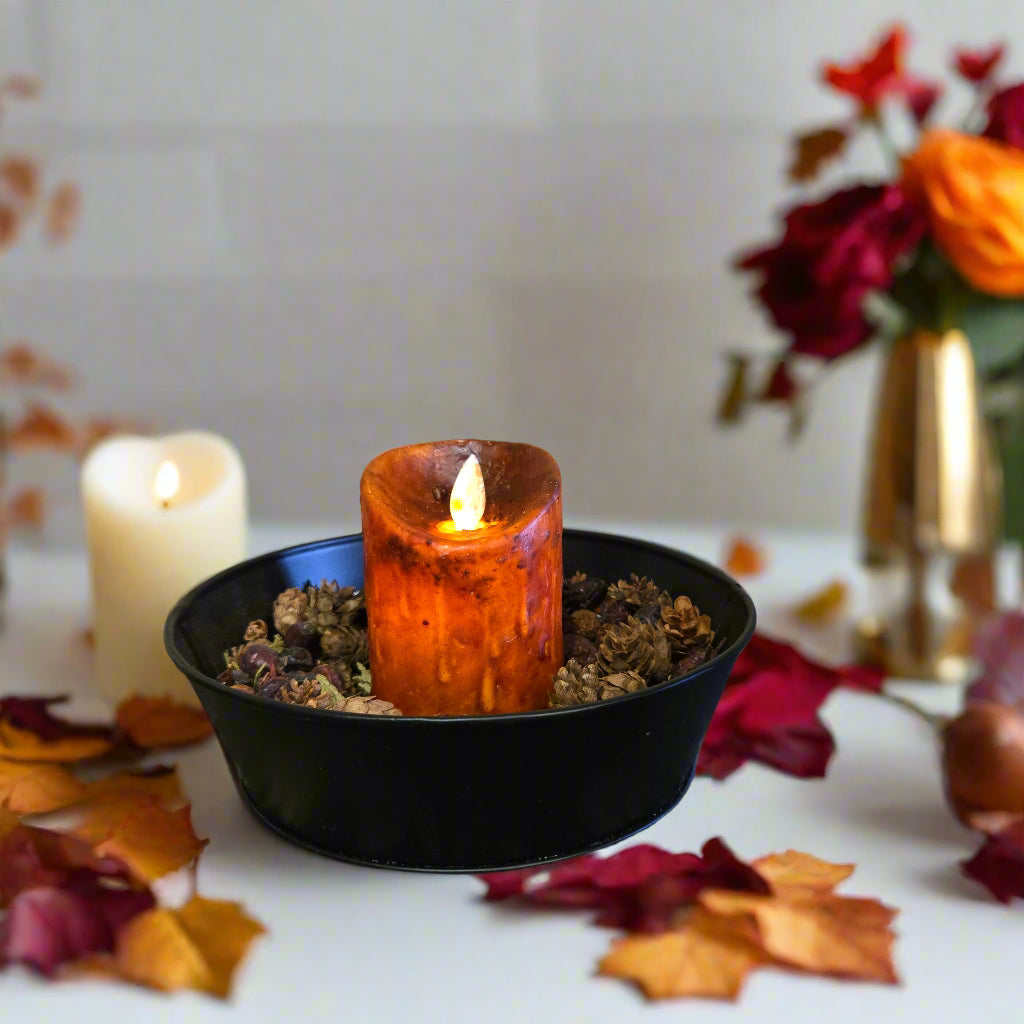 A burnt orange moving flame pillar candle placed in a black holder, situated on a textured surface with a wooden background.