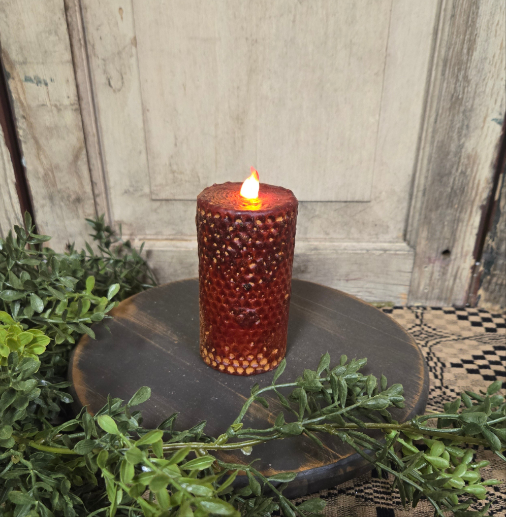 A battery-operated burgundy honeycomb-patterned LED pillar candle displaying a flame-shaped light, placed on a circular dark base surrounded by greenery.