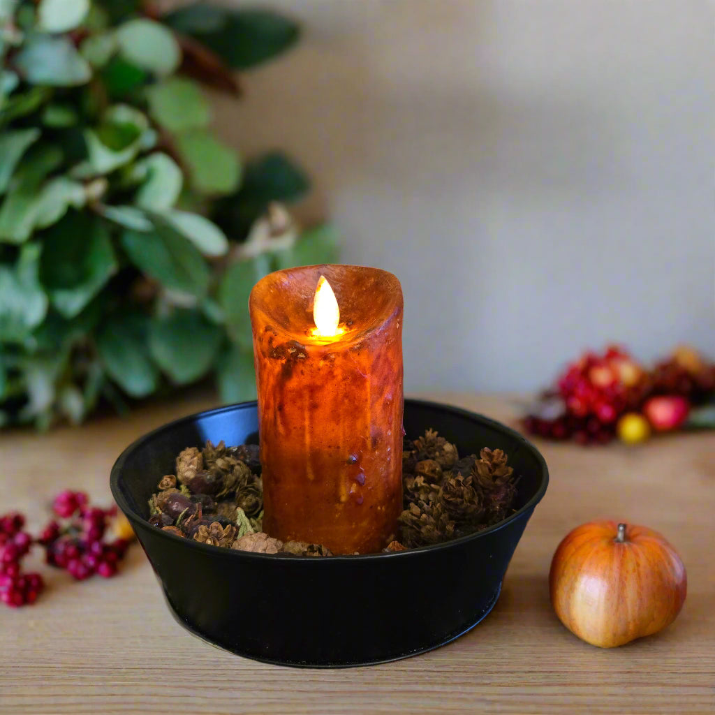 A burnt orange LED candle with a moving flame effect, placed in a black holder on a table with a lace tablecloth.