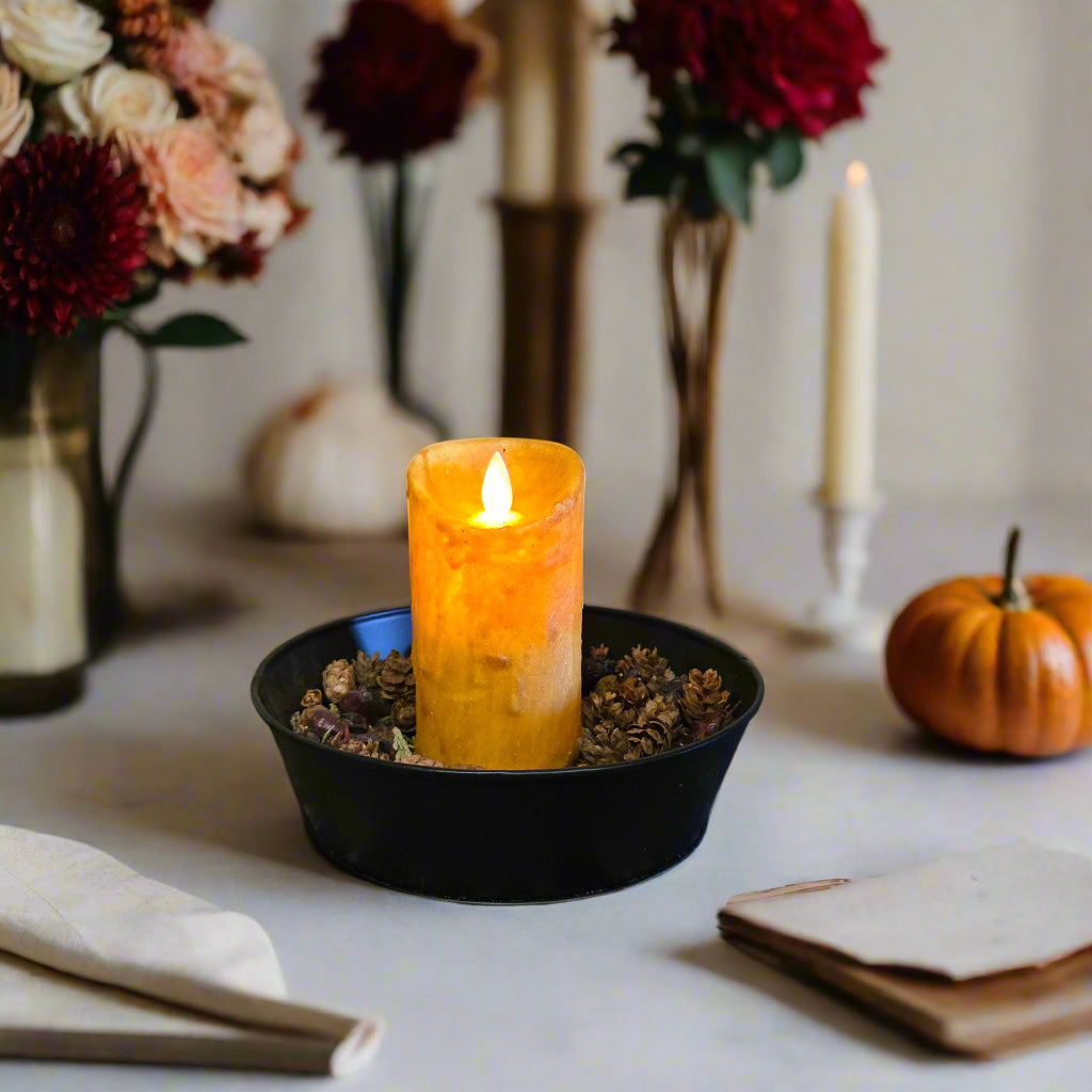 A 2" x 4" Moving Flame Pillar candle in a mustard color, placed on a black dish on a patterned fabric.