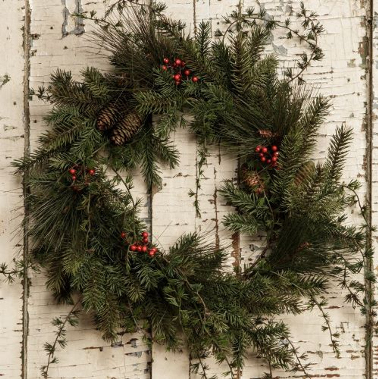 A round wreath made of mixed pine branches with red berries, displayed on a white wooden background.