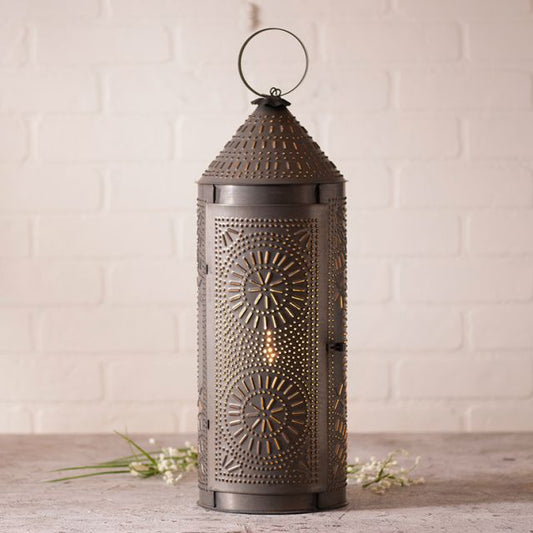A 22-inch high chimney lantern with a kettle black finish, featuring a geometric punch pattern, placed on a table with a white brick wall in the background and some decorative branches to the side.