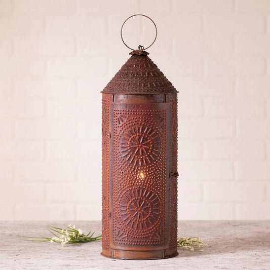 A 22-inch rustic tin chimney lantern with a punch pattern design, featuring a terracotta-like finish and a visible wick inside, placed on a grey surface with a white brick wall in the background.