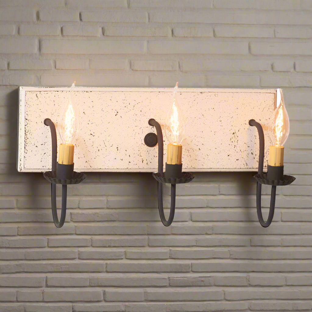 A vintage white three-light vanity light with black textured arms and a textured white painted wooden back, featuring three E12/Candelabra sockets.