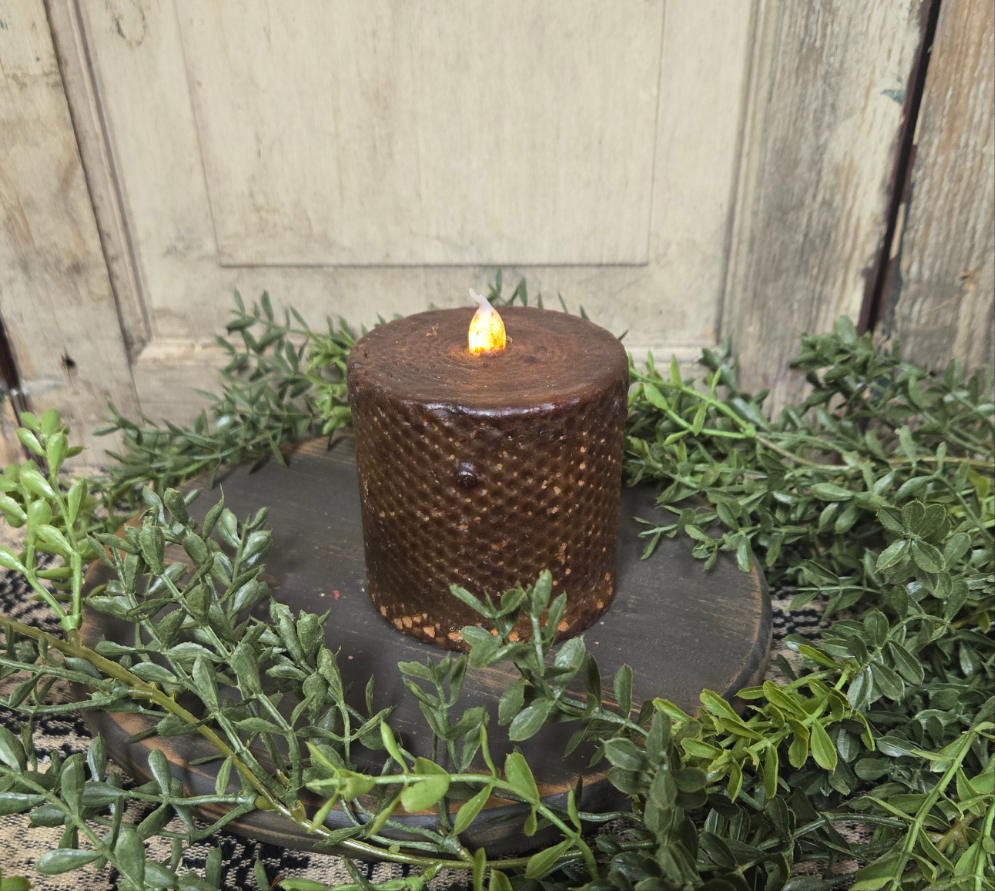 A black honeycomb-patterned pillar candle with a lit flame on top, surrounded by greenery and placed on a black round holder.