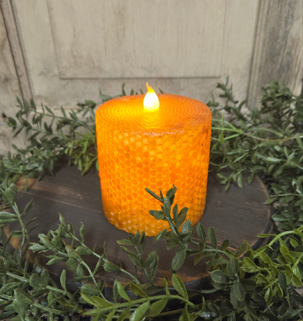 A battery-operated pillar candle with a black honeycomb-patterned wax coating, placed on a wooden holder, with a flame-shaped lightbulb that is lit, surrounded by green foliage.