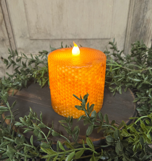 A battery-operated pillar candle with a black honeycomb-patterned wax coating, placed on a wooden holder, with a flame-shaped lightbulb that is lit, surrounded by green foliage.