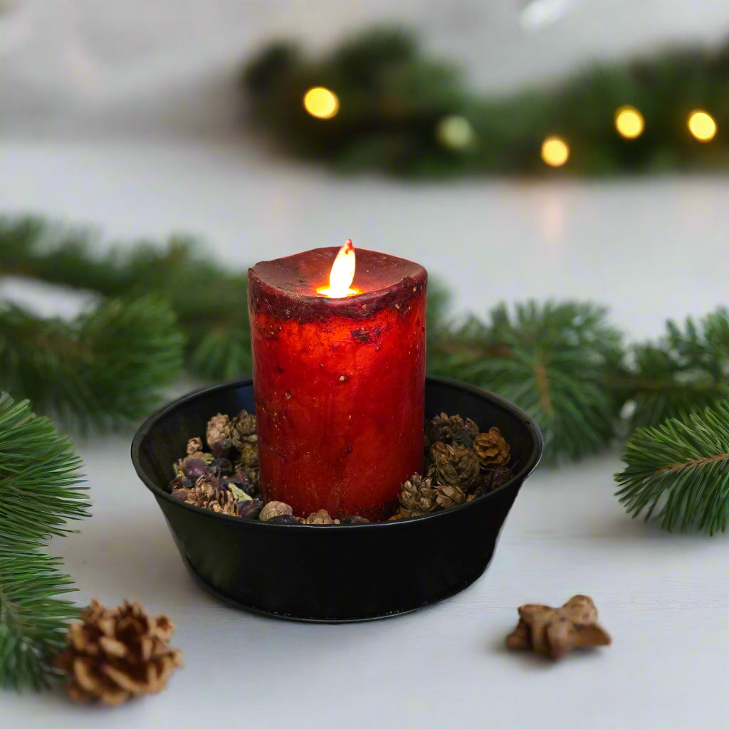 A synthetic moving flame candle in burgundy color appears to be burning, placed on a black dish on a patterned fabric.