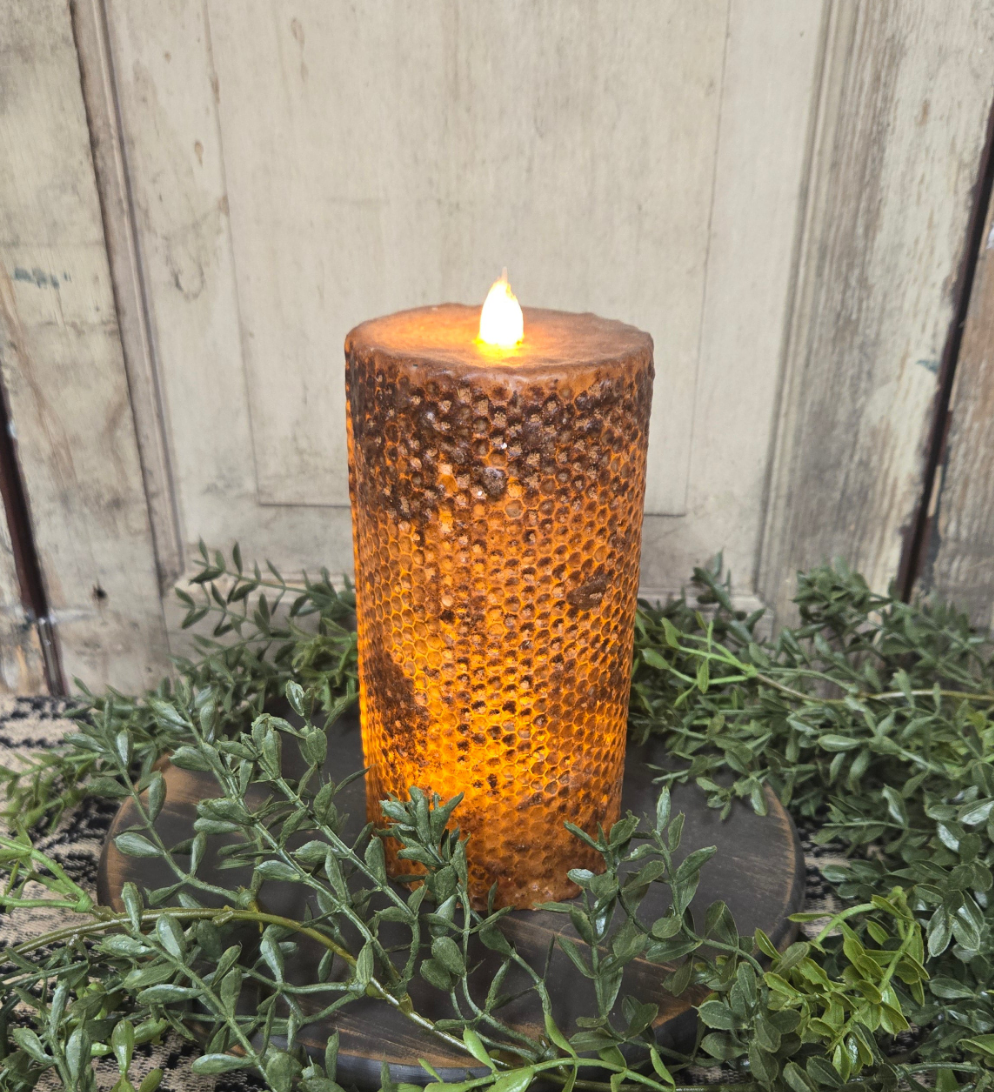 A brown honeycomb pillar candle with a lit flame on top, placed on a wooden stand surrounded by greenery.