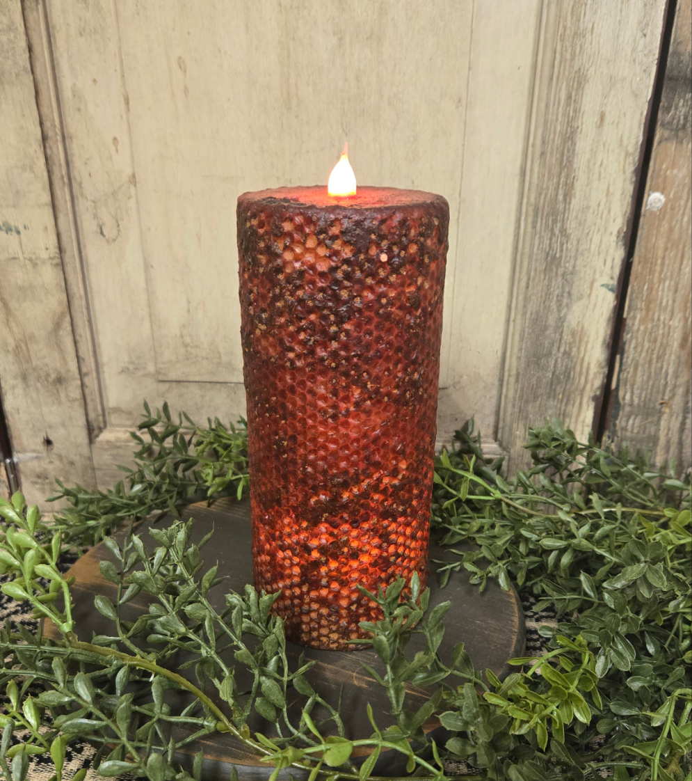 A burgundy wrapped honeycomb LED pillar candle with a lit flame on top, placed on a metal holder, surrounded by greenery.