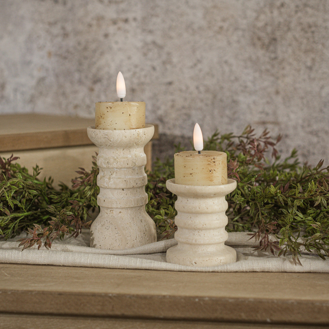 Two white cream tealight candles with a speckled flame effect, placed on a textured surface, surrounded by greenery.