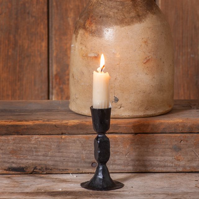 A black cast iron taper candle holder with a lit candle, placed on a wooden surface next to a beige candle.