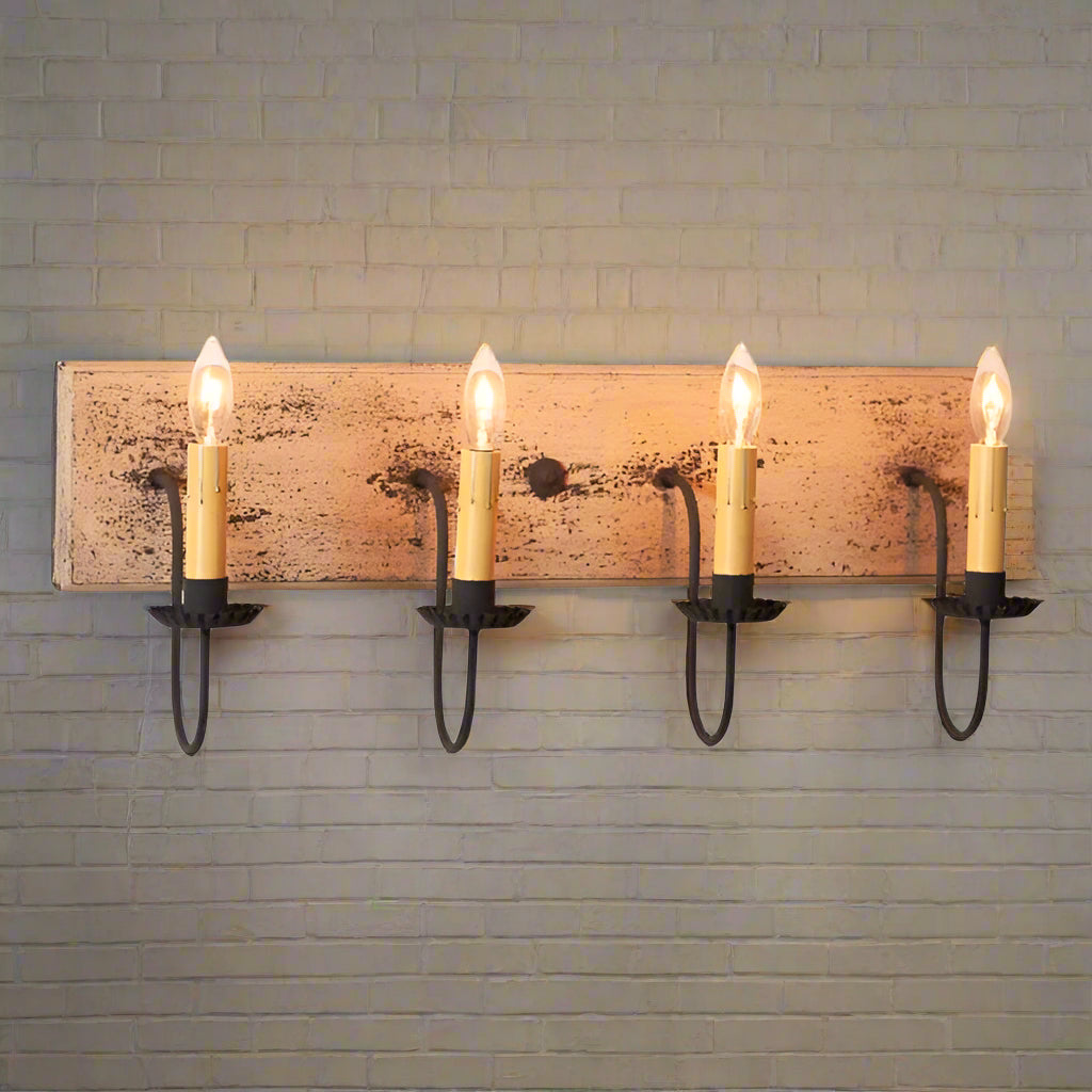 A four-light vanity light fixture with a heavily distressed painted finish in a buttermilk color, featuring textured arms with crimped pans and hanging from a wood panel.