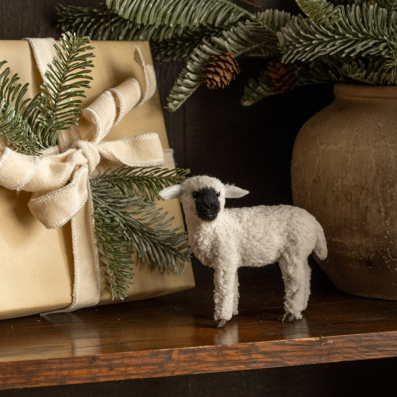 Small white sheep figurine next to a wrapped gift with greenery on a wooden surface.