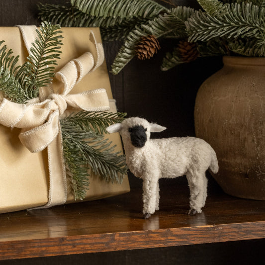 Small white sheep figurine next to a wrapped gift with greenery on a wooden surface.