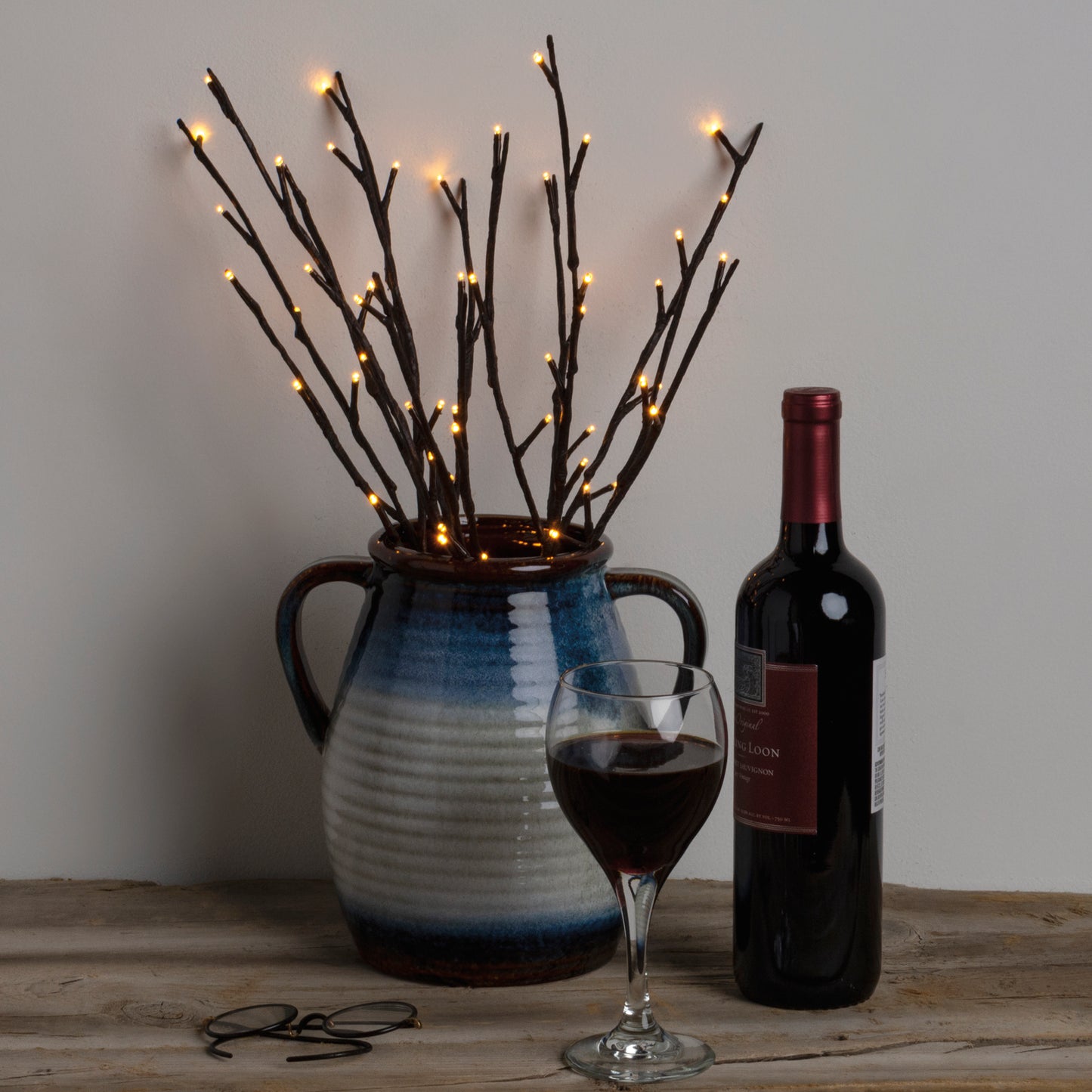 A battery-operated small willow twig tree with lights displayed in a blue and white vase next to a glass of red wine and a bottle.
