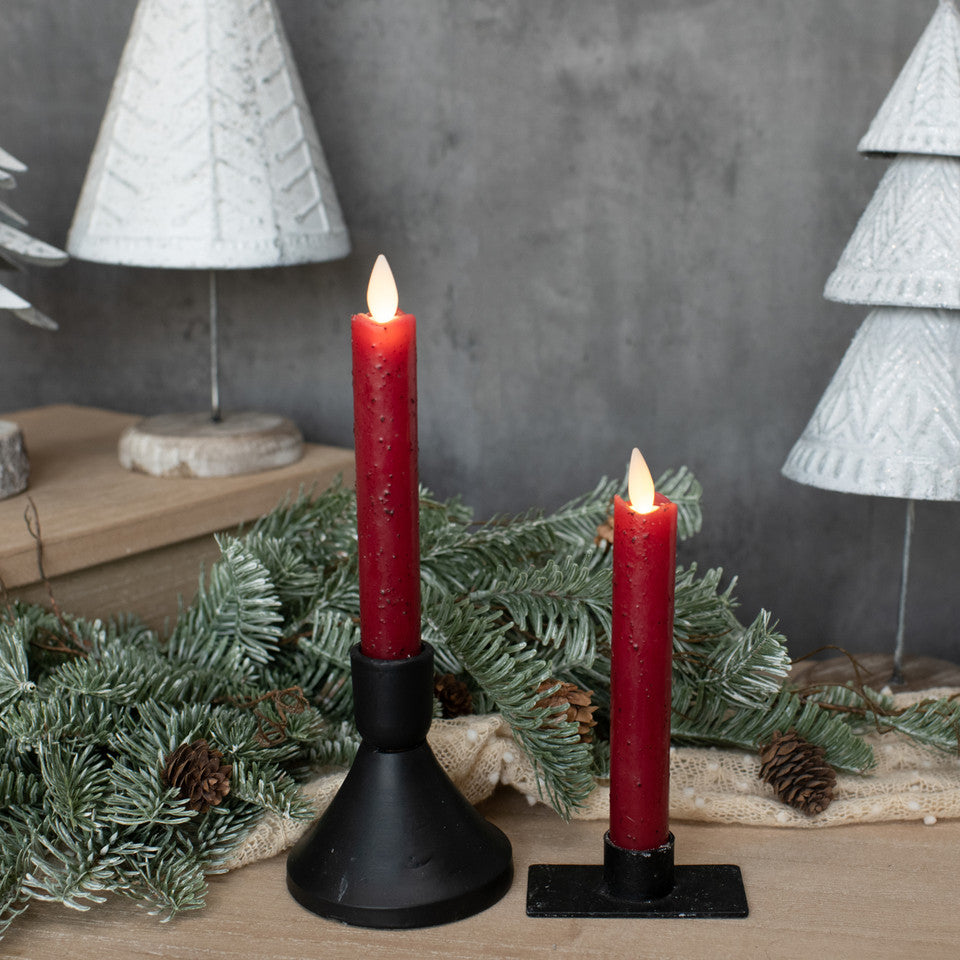 Two red candles in black holders with Christmas decorations on a wooden surface.