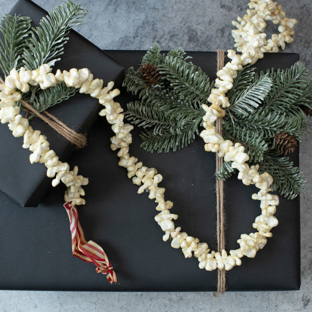 A 9-foot long popcorn garland made from artificial popcorn, displayed alongside green faux pine branches and red ribbon accents.