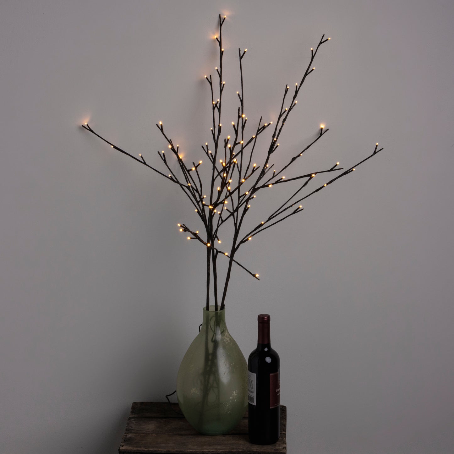 A large willow twig decorative lighting piece with numerous small lights, placed in a green vase, positioned on a surface next to a wine bottle and a glass.