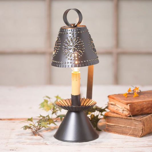 A smokey black accent lamp with a geometric pattern on the shade and a candelabra socket, placed on a wooden surface next to an open book and a decorative branch.