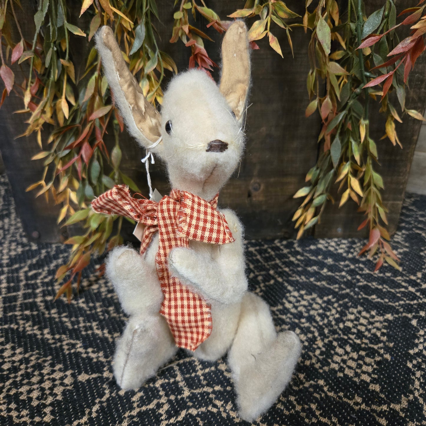 Small white bunny with a red checkered bow on a textured surface with greenery.