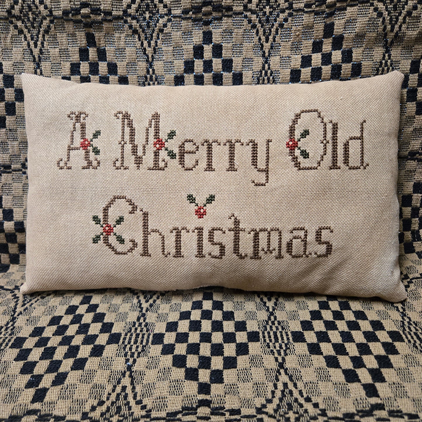 A rectangular beige linen pillow with 'A Merry Old Christmas' stitched in brown thread with small red and green holly accents, resting on a black and cream checkered patterned couch.