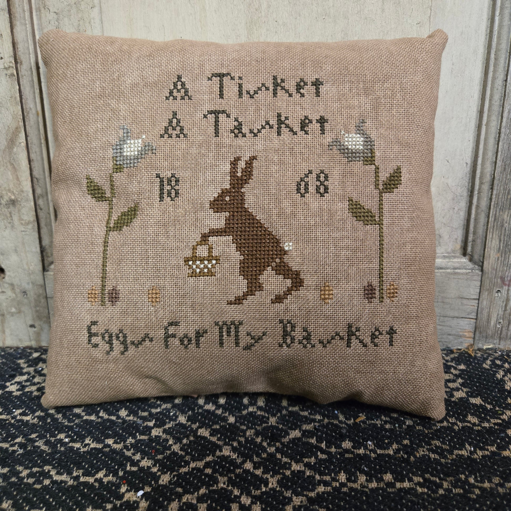 A rectangular cross stitch pillow with a bunny motif and the rhyme 'a tisket a tasket' embroidered on it, displayed on a textured surface.