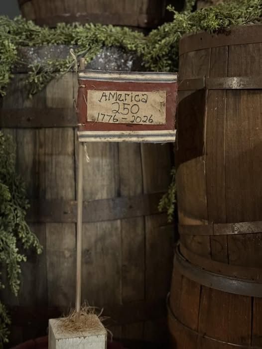 Wooden barrel with a sign celebrating America's 250th anniversary, surrounded by greenery.