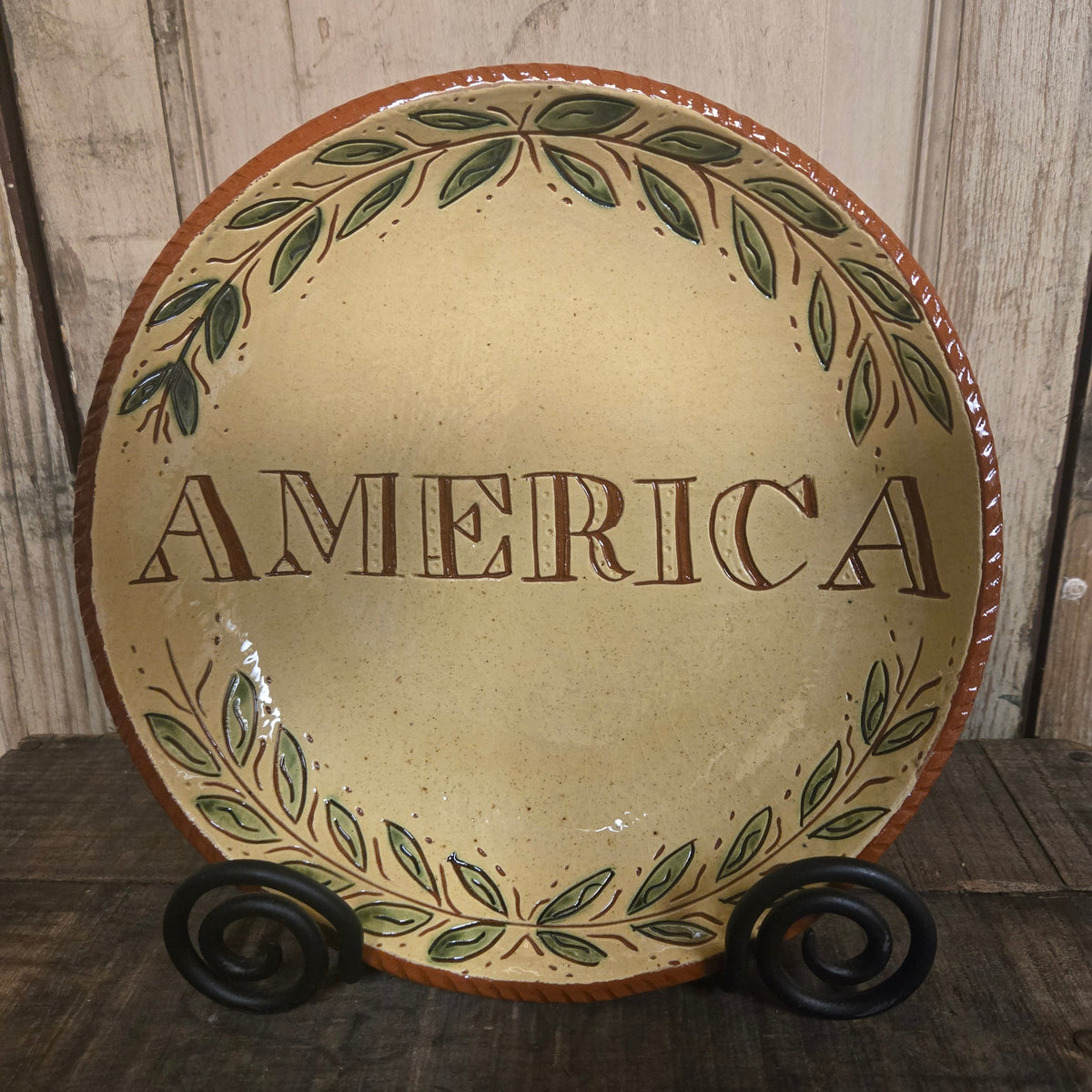 Cream-colored redware plate with “AMERICA” lettering, a green leafy border, and a rust-red rim, displayed on a black metal stand.