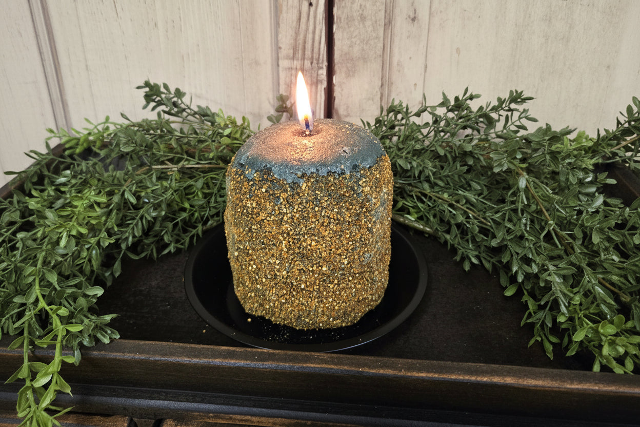A green and brown hearth candle with a primitive design, placed on a black dish, surrounded by greenery and set against a wooden background.