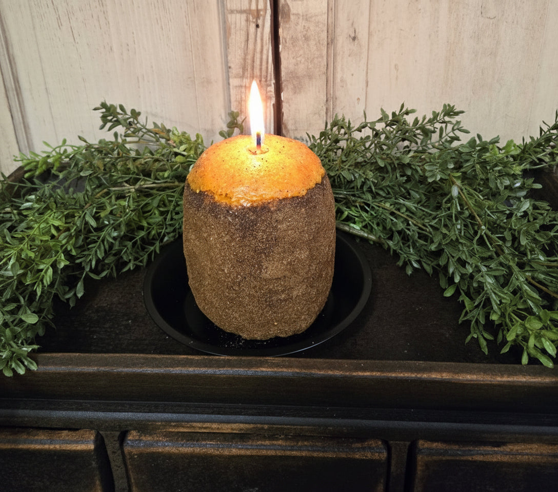A cylindrical gingerbread Hearth candle displayed on a hearth, surrounded by greenery. The candle has a brown exterior with a golden yellow top and is partially consumed, showing a visible wick.