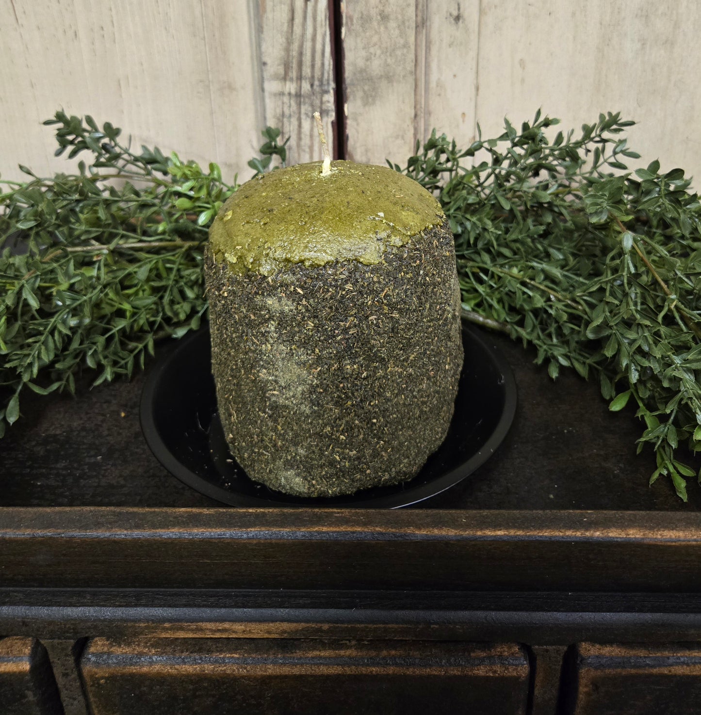 A green tomato-scented hearth candle placed on a black surface, surrounded by green foliage.