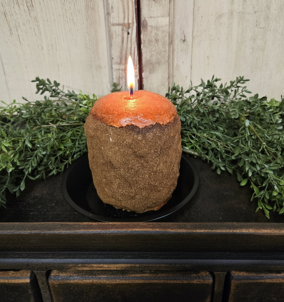 A vintage orange and clove scented hearth candle placed on a black dish, with a rustic wooden background and greenery around it.