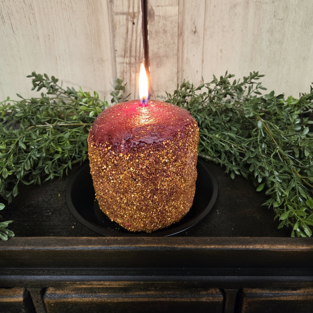 A spicy cranberry scented hearth candle placed on a black holder, with a rustic wooden background and greenery around it.
