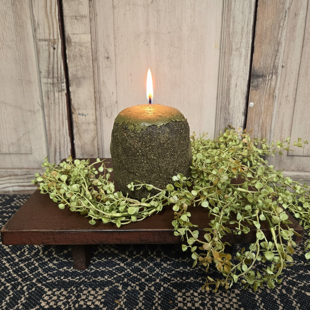 A green-colored hearth candle with a rough, textured surface, displayed on a wooden surface surrounded by a decorative green herb plant.