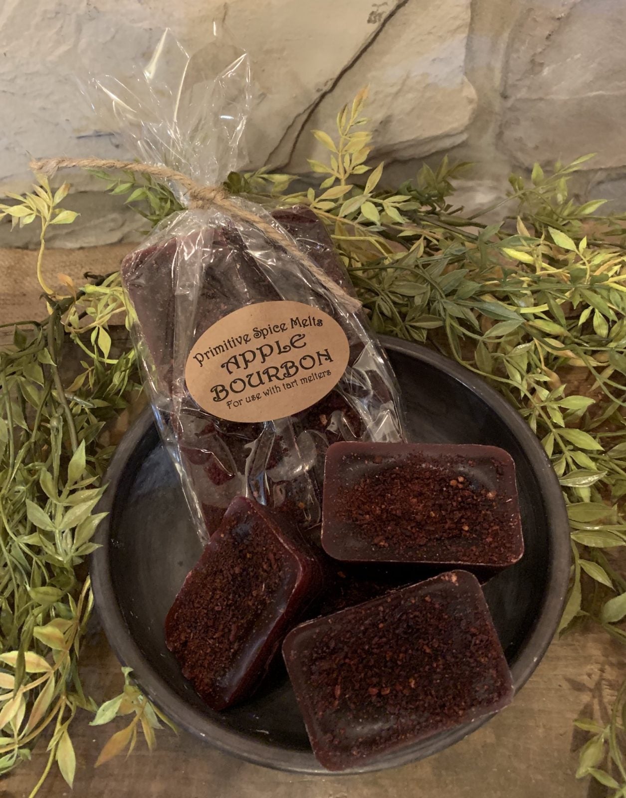A pack of burgundy wax melts labeled 'Apple Bourbon' placed on a dish with greenery in the background.
