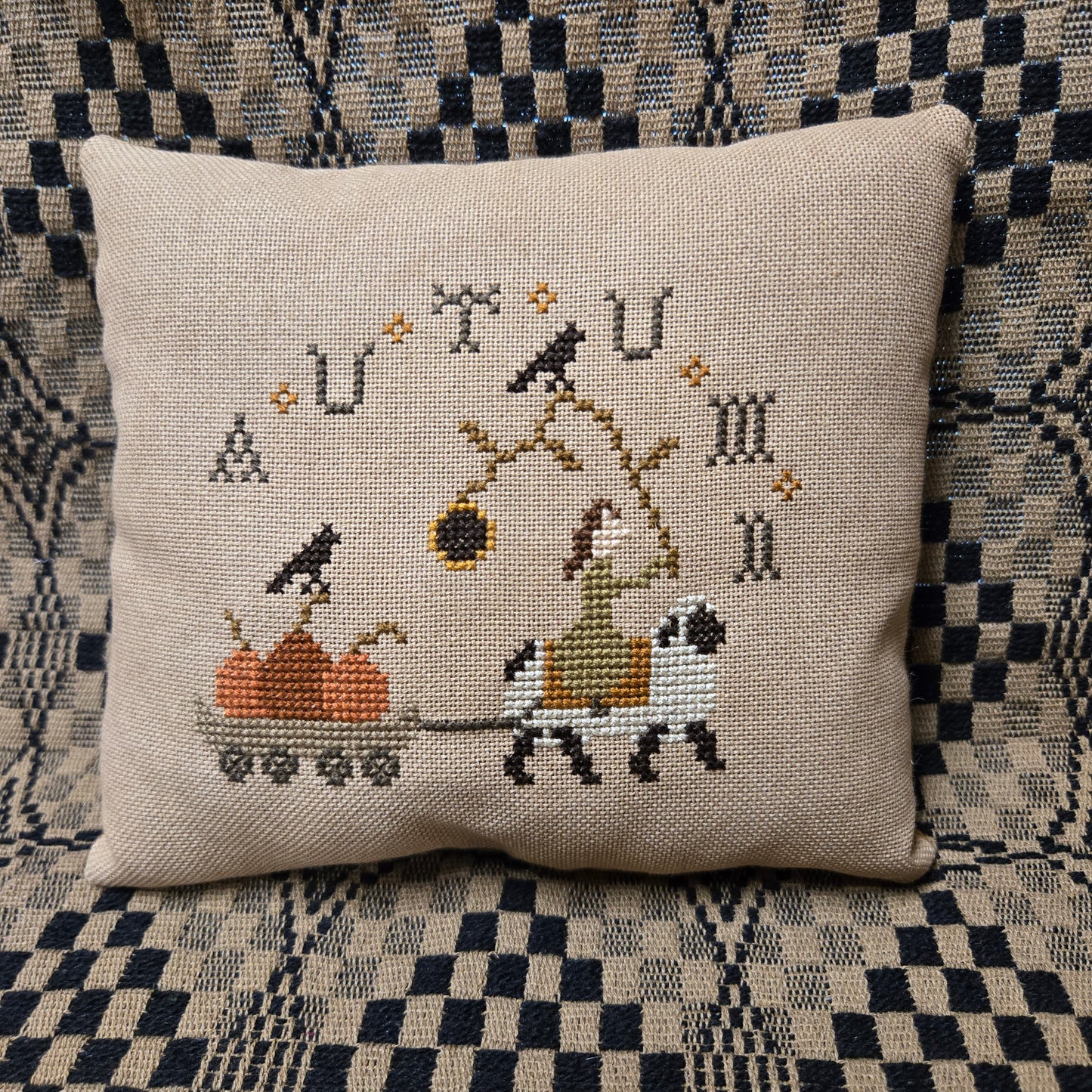 A square beige woven throw pillow featuring a cross-stitched autumn-themed design with a sheep, cart, and pumpkins displayed on a black and white checkered patterned couch.
